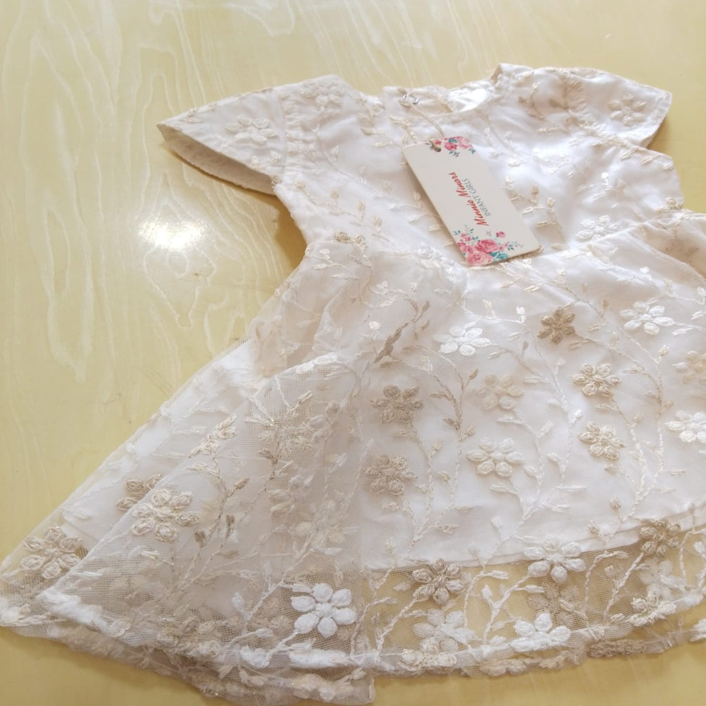 A cream-colored lace fan with embroidered floral details, laid flat on a light beige surface. A small paper tag with a floral design is attached near the center.