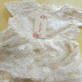 An ivory white lace baby bodysuit with floral embroidery and a matching paper tag, laid flat on a light yellow surface.