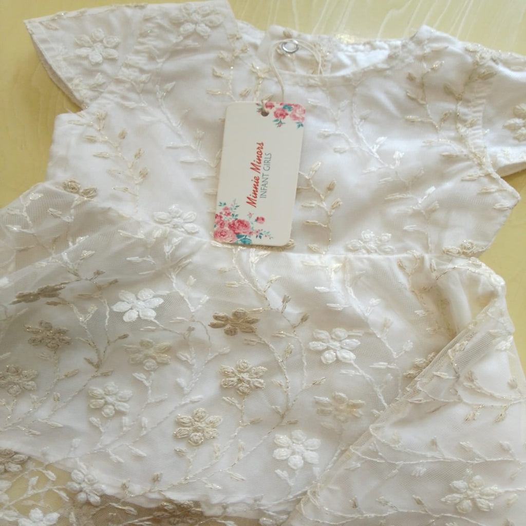 An ivory white lace baby bodysuit with floral embroidery and a matching paper tag, laid flat on a light yellow surface.