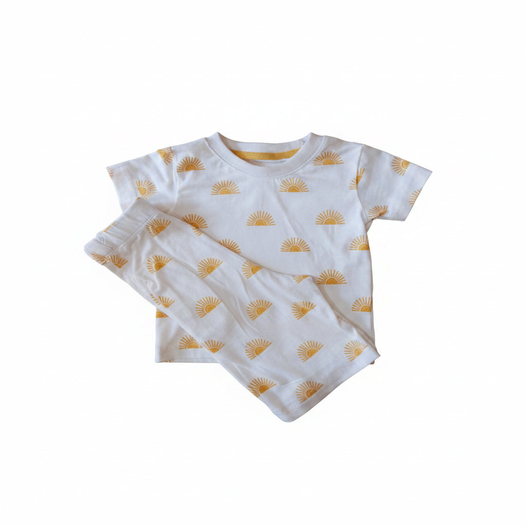 A two-piece baby pajama set consisting of a short-sleeve white top and matching pants, both printed with a small yellow sunburst pattern, laid flat on a light wood surface.