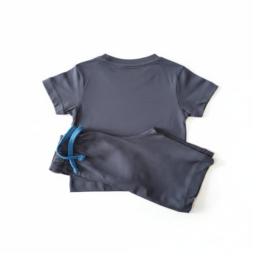 A two-piece kids' pajama set consisting of a short-sleeve t-shirt and matching shorts, laid flat on a light wood floor. The set is dark gray with a bright blue drawstring on the shorts.